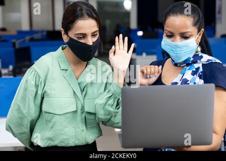Porträt von zwei jungen indischen Geschäftsfrauen tragen covid Schutzmaske, winkende Hand an Laptop, Videokonferenz, Client-Meeting, Büroumgebung. Stockfoto
