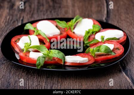 Selektiver Fokus. Caprese Salat mit Tomatenscheiben, Mozzarella und Basilikum auf einem schwarzen Teller auf einem Holzhintergrund. Stockfoto