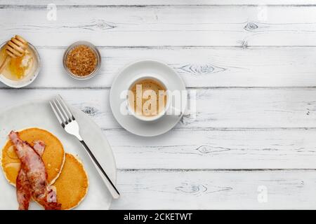 Ein Teller mit Speck und Pfannkuchen, mit Honig beträufelt, eine Tasse Kaffee und eine Schüssel mit Zucker, auf einem rustikalen weißen Holzhintergrund mit Platz für Kopie Stockfoto