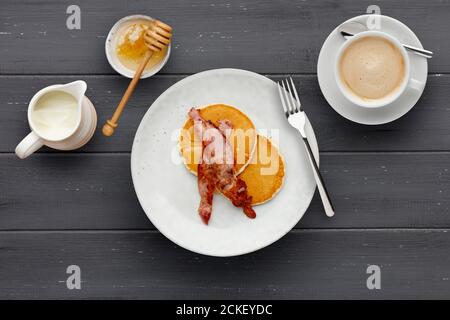 Ein Teller mit Speck und Pfannkuchen, mit Honig beträufelt, eine Tasse Kaffee und eine Kanne Sahne auf einem dunklen Holzhintergrund Stockfoto