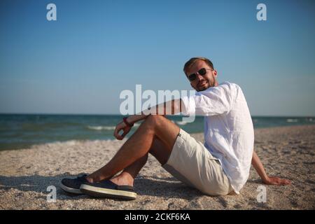 Junger Mann in Sonnenbrille sitzt und lächelt am Strand vor Meer und Himmel Hintergrund. Stockfoto