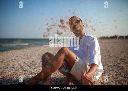 Junger Mann in Sonnenbrille sitzt am Strand, hat Spaß, lacht und wirft Sand gegen Meer und Himmel Hintergrund. Stockfoto