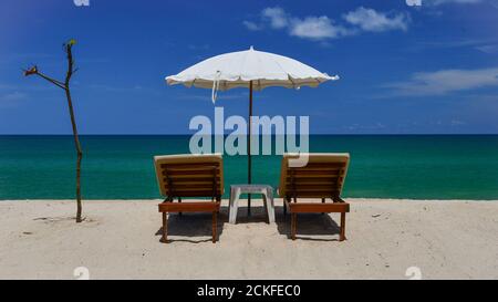 Rückansicht von einem Paar Holzslatten-Sonnenliegen an einem weißen tropischen Sandstrand unter einem weißen Sonnenschirm. Stockfoto