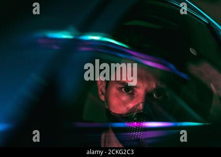 Ein junger Mann in einer Gesichtsmaske, der ein Auto fährt In der Nacht Stadt mit Neonlicht, die von der Glas seines Autos Stockfoto