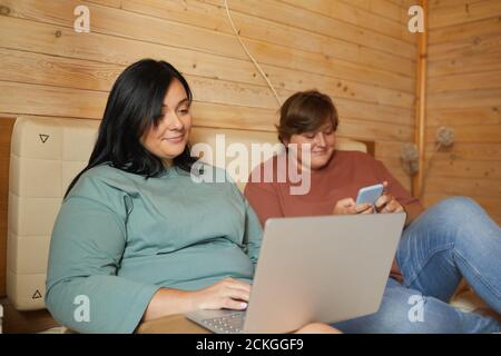 Junge übergewichtige Frau liegt auf dem Bett und mit Laptop mit Ihre Freundin mit dem Handy ruhten sie zusammen Stockfoto