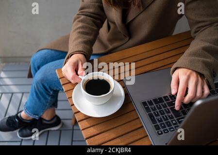 Frau in Wollmantel, die im Straßencafé mit Laptop arbeitet. Tasse Kaffee auf Holztisch. Stockfoto