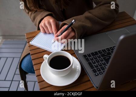 Frau in Wollmantel, die im Straßencafé mit Laptop arbeitet. Tasse Kaffee auf Holztisch. Schreiben im Notizblock Stockfoto