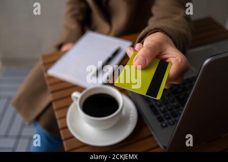 Frau in Wollmantel, die mit Karte im Straßencafé bezahlt. Arbeiten mit Laptop, Notizblock und Kaffee Stockfoto