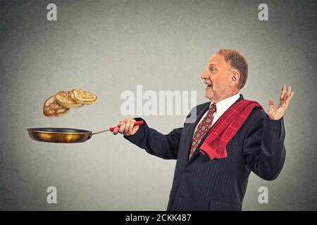 Glücklich lächelnder älterer Mann, der Pfannkuchen auf Bratpfanne wirft, isoliert auf grauem Wandhintergrund. Positive Gesichtsausdruck Emotion, Küche Spaß Konzept Stockfoto