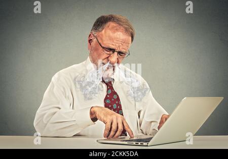 Verwirrt aussehende ältere ältere Mann mit Brille am Tisch sitzen Arbeiten mit dem Tippen auf einem Laptop-Computer Stockfoto