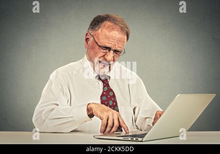 Verwirrt aussehende ältere ältere Mann mit Brille am Tisch sitzen Arbeiten mit dem Tippen auf einem Laptop-Computer Stockfoto