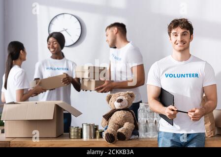 Selektiver Fokus der Freiwilligen hält Zwischenablage und Stift in der Nähe multiethnisch Menschen mit Paketen in Charity-Center Stockfoto