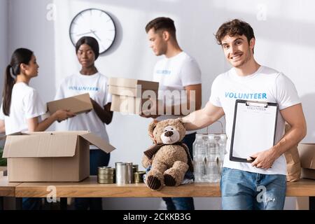 Selektiver Fokus der Freiwilligen hält Zwischenablage und weiches Spielzeug in der Nähe Multiethnische Menschen in Wohltätigkeitszentrum Stockfoto