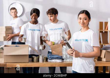 Selektiver Fokus der asiatischen Freiwilligen hält Zwischenablage und Blick auf Kamera in der Nähe von multiethnischen Menschen und Spenden in Wohltätigkeitszentrum Stockfoto