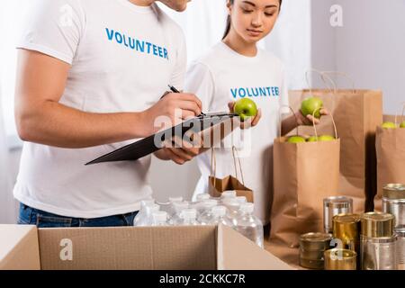 Selektiver Fokus des Mannes, der auf Zwischenablage schreibt, nahe asiatischer Freiwilliger Äpfel und Pakete im Charity Center halten Stockfoto