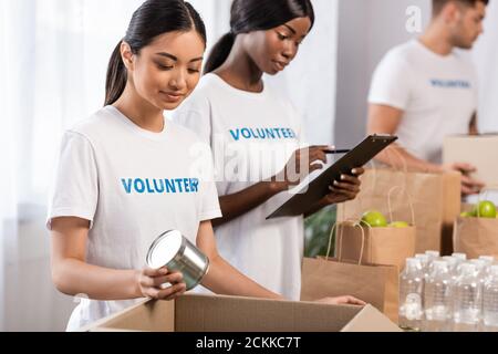 Selektiver Fokus der asiatischen Freiwilligen hält Zinndose in der Nähe von african american Frau schriftlich auf Zwischenablage in Charity-Center Stockfoto