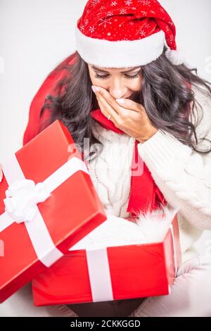 Mädchen in einem Weihnachtshut öffnet ein Weihnachtsgeschenk und hält Hand vor ihrem Mund vor der Überraschung. Stockfoto