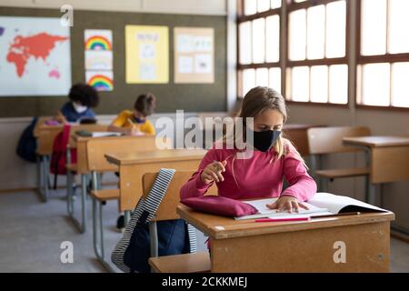 Mädchen trägt Gesichtsmaske studieren, während sie auf ihrem Schreibtisch sitzen In der Schule Stockfoto