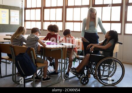 Lehrerin mit digitalem Tablet im Gespräch mit einem deaktivierten Mädchen In der Schule in ihrem Rollstuhl sitzend Stockfoto