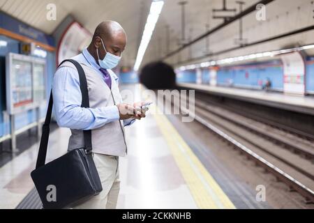 Schwarzer Mann in einer Gesichtsmaske mit seinem Smartphone in einer U-Bahn-Station. Sichere Reise und neue Normalität während der Covid-19 Pandemie. Stockfoto