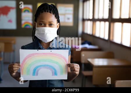 Portrait des Mädchens trägt Gesichtsmaske hält eine Regenbogenzeichnung In der Schule Stockfoto