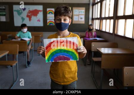 Portrait des Jungen trägt Gesichtsmaske hält eine Regenbogenzeichnung In der Schule Stockfoto