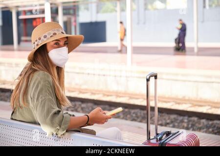 Die junge Frau trägt eine Gesichtsmaske und benutzt ihr Smartphone, während sie am Bahnhof auf den Zug wartet. Reisekonzept. Stockfoto