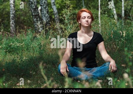 Mädchen Teenager meditiert im Park. Rothaarige Frau, die auf Gras sitzt. Freiheit und freudiges Konzept mit Kopierraum. Stockfoto