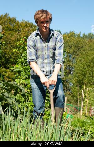Junger Mann mit Gummistiefeln, der an einem sonnigen Frühlingstag in einem Gemüsegarten graben. Stockfoto
