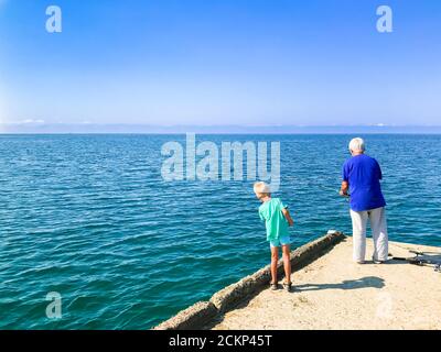 Der kleine Junge sieht einem alten Mann beim Fischen im Meer zu, während er auf dem Pier steht. Stockfoto