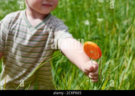 Hand eines kleinen Jungen mit einem Lollipop in Form einer geschnittenen Orange auf einem Hintergrund von Gras. Stockfoto