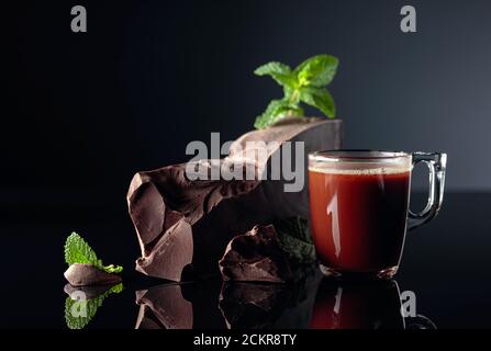 Schokolade und Minze. Stücke von dunkler Bitterschokolade und eine Glastasse mit heißer Schokolade auf schwarzem reflektierendem Hintergrund. Speicherplatz kopieren. Stockfoto