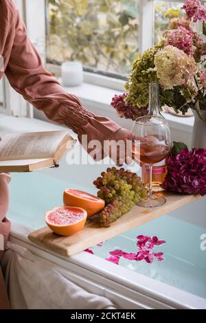 Badewanne mit Blütenblättern, Grapefruitscheiben, Trauben und Frauenhand mit einem Glas Wein und einem Buch. Bio-Spa Entspannung präparatio Stockfoto