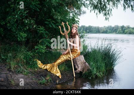 Fantasy Frau echte Meerjungfrau mit Dreizack Mythos Göttin des Meeres mit goldenen Schwanz sitzt in Sonnenuntergang auf Felsen.. Gold Haar Krone Muscheln Perlen Schmuck. Merma Stockfoto