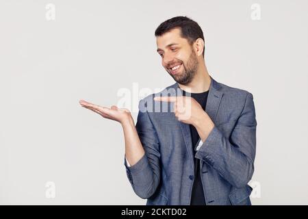 Ein junger Geschäftsmann mit Bart in einer Jacke blickt auf die Handfläche, zeigt mit der Hand und zeigt mit dem Finger. Porträt eines Mannes auf grauem Hintergrund. Stockfoto