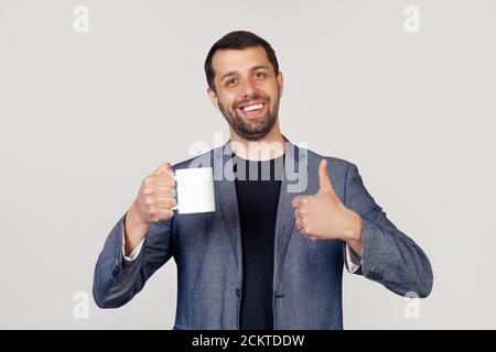 Ein junger Geschäftsmann mit einem Lächeln, ein Mann mit Bart in einer Jacke, eine Tasse Kaffee in der Hand und eine Geste mit dem Daumen nach oben, ein Mann mit einem glücklichen Gesicht. Porträt eines Mannes auf grauem Hintergrund. Stockfoto