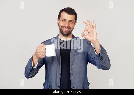 Junger Geschäftsmann mit einem Lächeln, ein Mann mit einem Bart in einer Jacke, hält eine Tasse Kaffee und zeigt die Geste alles ist in Ordnung, ein Mann mit einem glücklichen Gesicht. Porträt eines Mannes auf grauem Hintergrund. Stockfoto