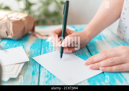 Eine Frau schreibt auf einer leeren Postkarte. Hände, die einen Stift in Nahaufnahme halten. Blauer Holztisch mit einem Paket im Hintergrund. Das Konzept der Post entsprach Stockfoto