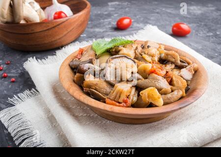 Gebratene Austernpilze mit Tomaten in Holzplatte auf schwarzem Beton Hintergrund. Seitenansicht, Nahaufnahme Stockfoto