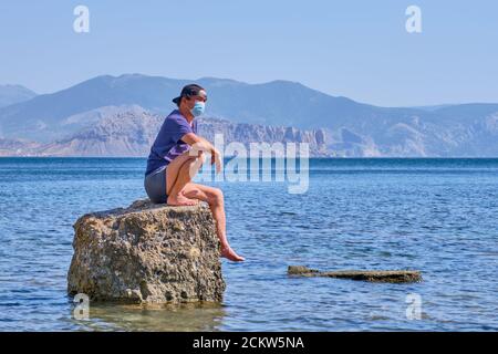 Senior Asian Mann in Gesichtsmaske, Shorts, T-Shirt und Baseball-Cap sitzt allein auf einem Stein in einer Meeresbucht vor einem Hintergrund von Bergen Stockfoto
