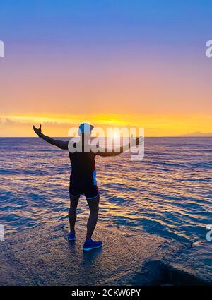 Guy steht am Ufer des Piers mit den Händen nach oben und schaut überraschend den Sonnenuntergang im Meer mit einem wundervollen orangefarbenen Himmel an Stockfoto