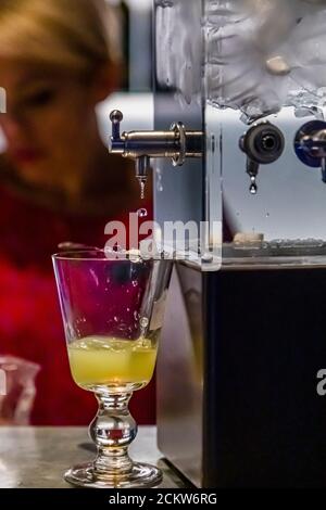 Gelb-grüne Absinthe fließt aus dem Hahn eines Wasserbrunnens in ein Glas im Maison de l'Absinthe in Môtiers; eine Figur in Rot steht verschwommen im Hintergrund. Das Wasserritual mit Zuckerwürfel und Löffel trübt die Absinthe allmählich milchig. Das Val-de-Travers gilt seit 1797 als Geburtsort der Absinthe und liegt im Kanton Neuenburg, Schweiz. Stockfoto