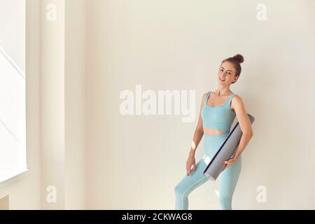 Yoga. Junge weibliche Turnerin mit Übungsmatte in der Hand, die neben der Wand des Fitnessraums steht und die Kamera anschaut Stockfoto