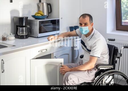 Lächelnder Junger Behinderter Mann, Der In Der Küche Auf Dem Rollstuhl Sitzt. Junger Mann mit Gesichtsmaske sitzt vor der Küche. Konzentrieren Sie sich auf sein Gesicht. Stockfoto