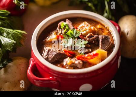 Schüssel Gulaschsuppe mit Fleisch, Kartoffeln und Chilis neben rohen Zutaten für den Eintopf Stockfoto
