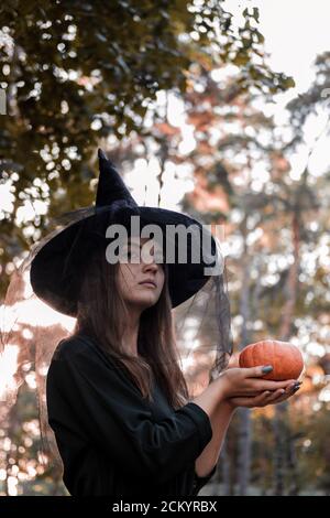 Junge Frau in dunklem Kleid und Hexenhut hält einen Kürbis in den Händen. Halloween Party Kostüm. Wald, Park mit Herbstbäumen Stockfoto