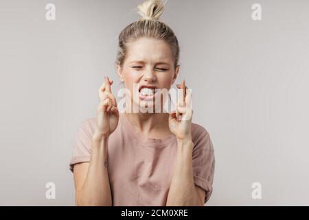 Studio isoliert Porträt von attraktiven blonde Studentin mit Haar-Brötchen, halten die Daumen gedrückt und die Augen geschlossen, in der Hoffnung auf Glück, bevor Sie gehen Stockfoto