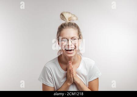 Fröhliche blonde junge Frau trägt stilvolle Haarknoten und Brille lachend Kamera, klammernde Hände zusammen, beten, um zu stoppen lachen sie, isol Stockfoto