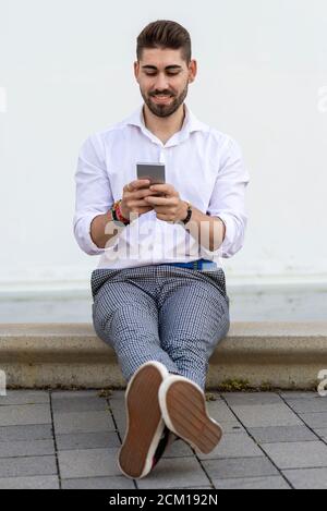 Junger bärtiger Mann sitzt an einer weißen Wand mit dem Mobiltelefon Stockfoto