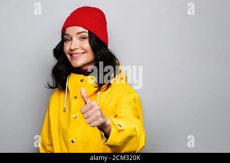 Glücklich attraktive Brünette Frau in gelben Herbst Regenmantel und rote Wollmütze, zeigt Daumen nach oben Geste, lächelnd, akzeptieren guten Plan, Genehmigung tun Position Stockfoto